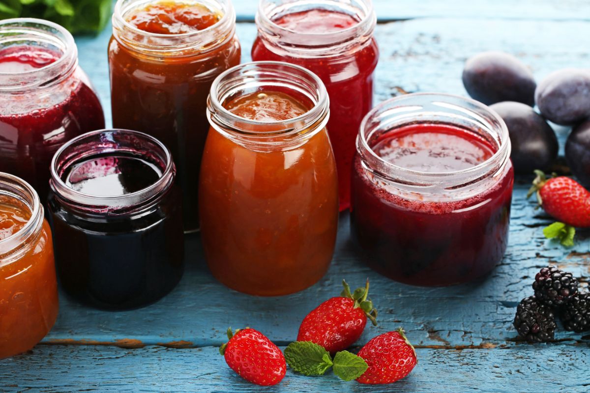 Different varieties of jams in  glass jars on a wooden table with scattered fruits around.