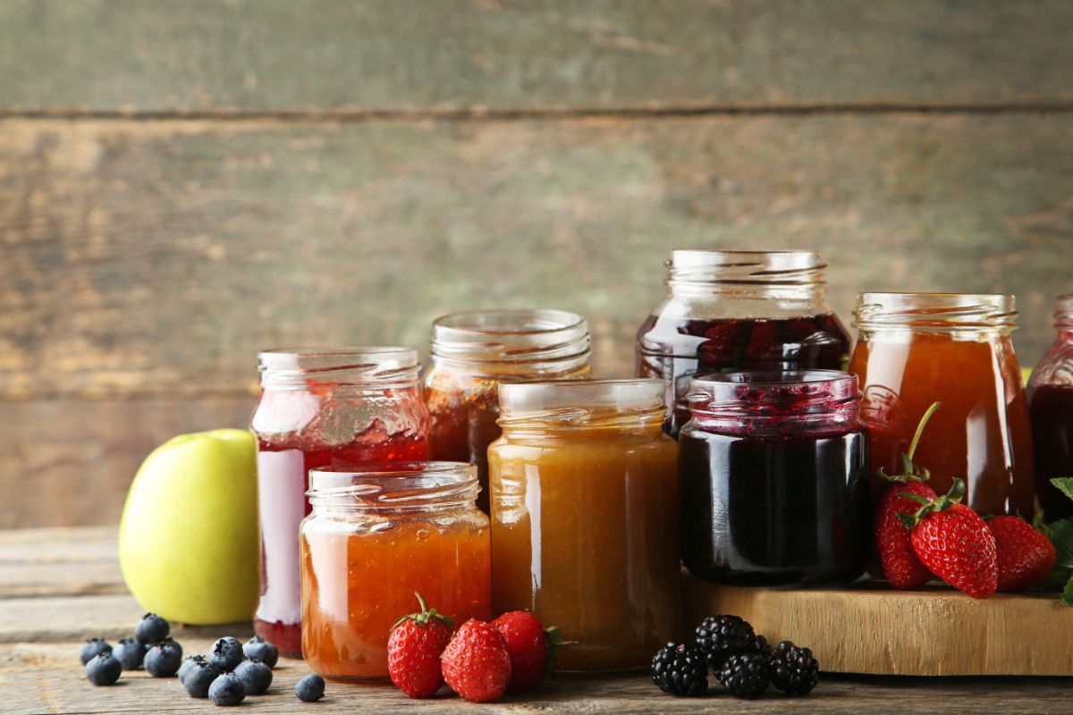 Different varieties of jams in glass jars on a table with scattered fruits aorund.