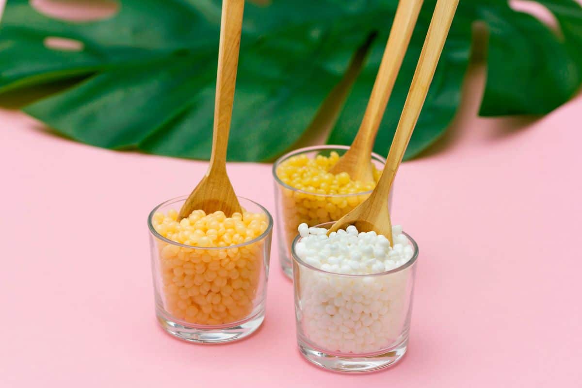 Wax beads in glass container with wooden spatulas on a pink table.