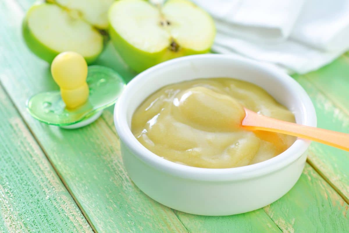 A white bowl full of baby food with a spoon, a soother, and sliced apples on a table.
