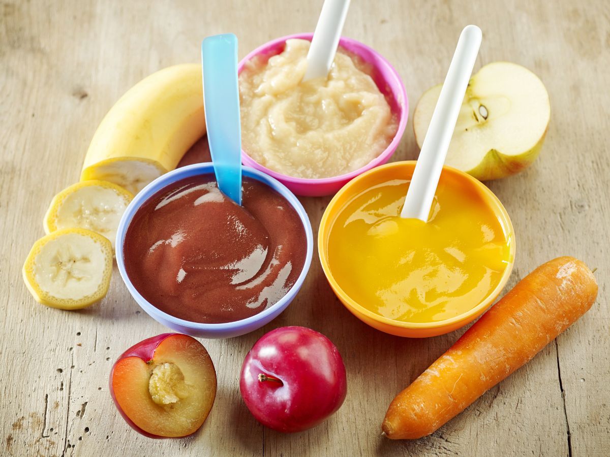 Three bowls of baby foods with spoons and sliced fruits and vegetables on a table.