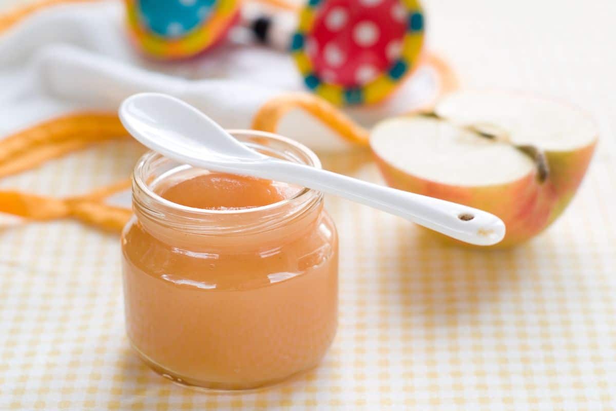 A glass jar of baby food with a white spoon on a table with a sliced apple.