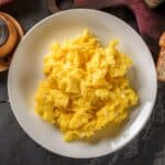A white plate full of scrambled eggs on a wooden table.