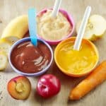 Three bowls of baby foods with spoons and sliced fruits and vegetables on a table.