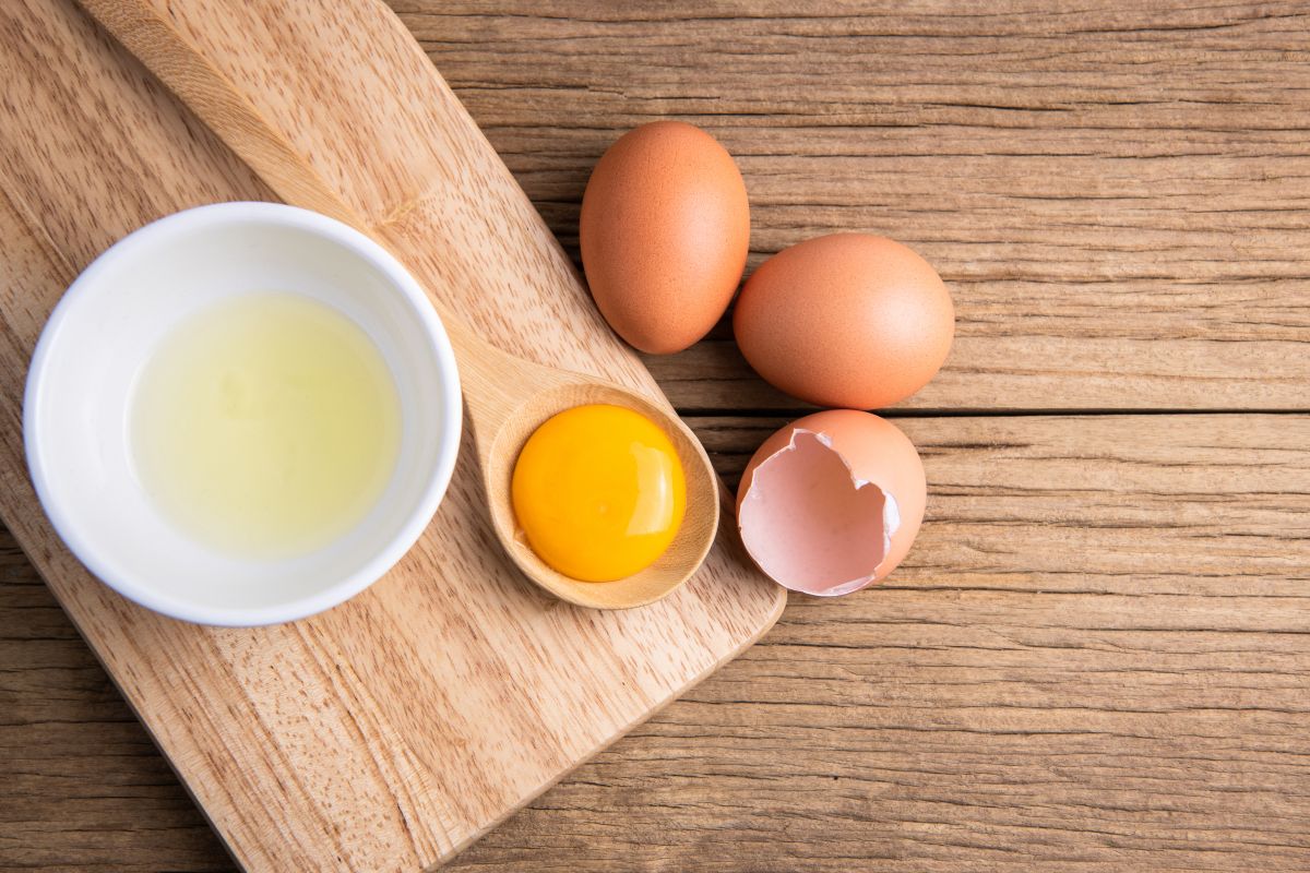 A white bowl of egg whites with a wooden spoon with folk on a wooden cutting board next to two whole eggs and an eggshell.