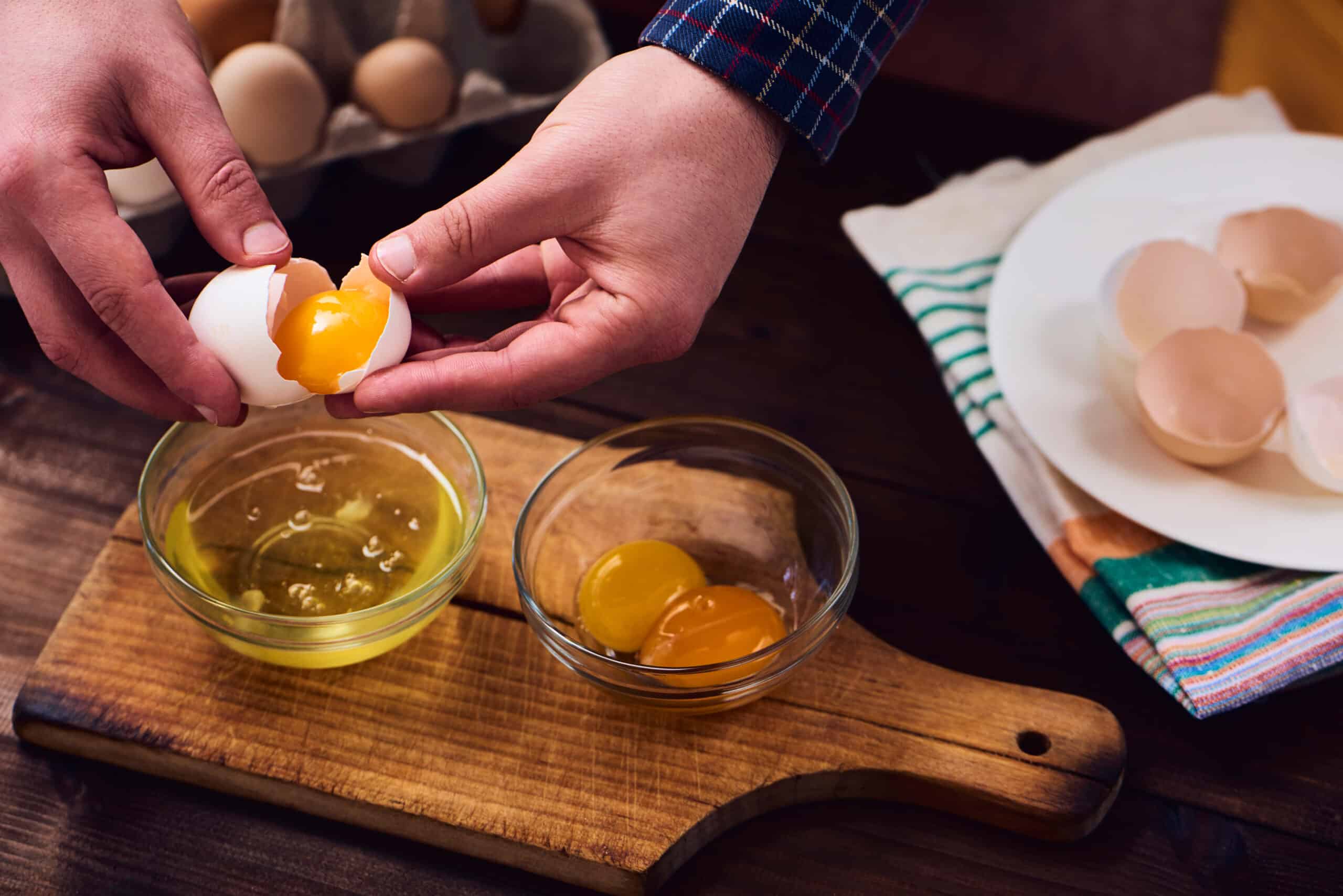 A man breaks up an egg over two small glass bowl on a wooden cutting board.