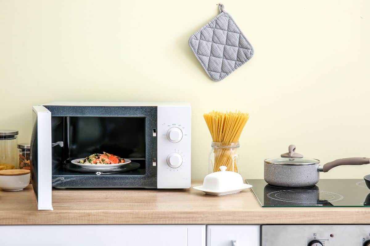 A microwave with food on a plate on the kitchen counter with a pot, and many cooking ingredients and kitchenware.