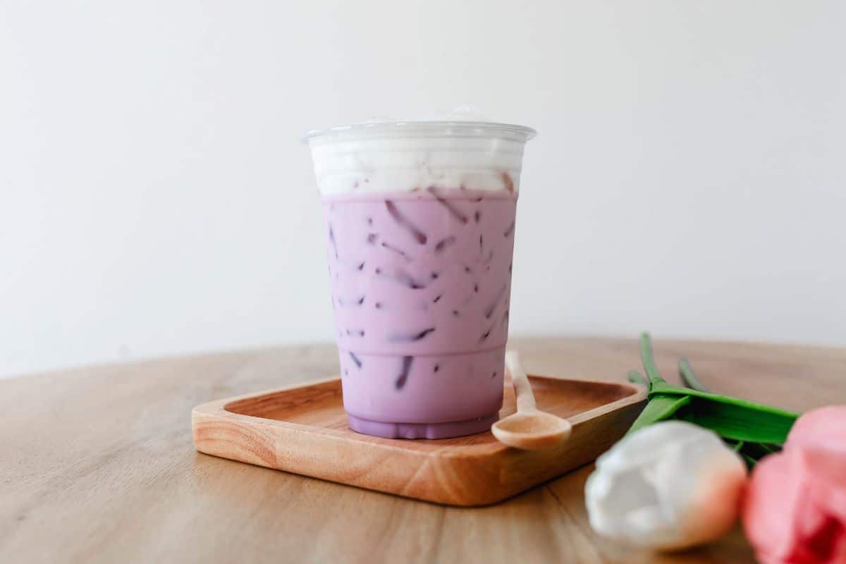 A plastic cup full of purple tea on a small wooden tray with a spoon on a table with roses.