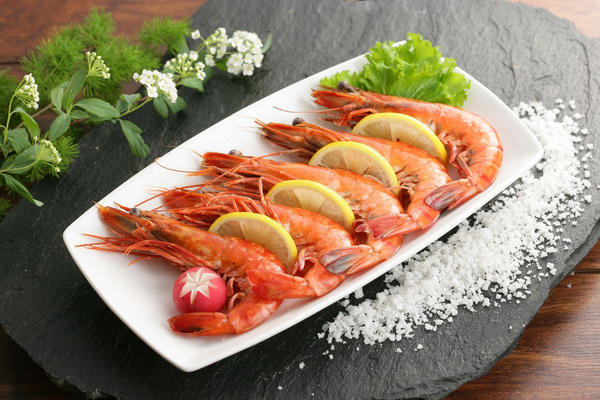 A white tray full of shrimp, slices of lemons, and vegetables on a black board with salt and herbs.