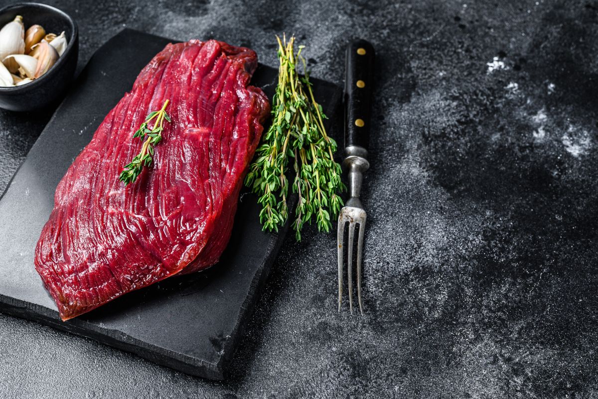 A raw steak on a black board with herbs, a for,k and a bowl of garlic on a black table.