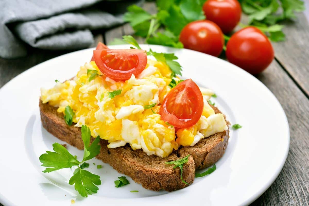 Scrambled eggs on a slice of bread with tomatoes, and herbs on a white plate on a wooden table.