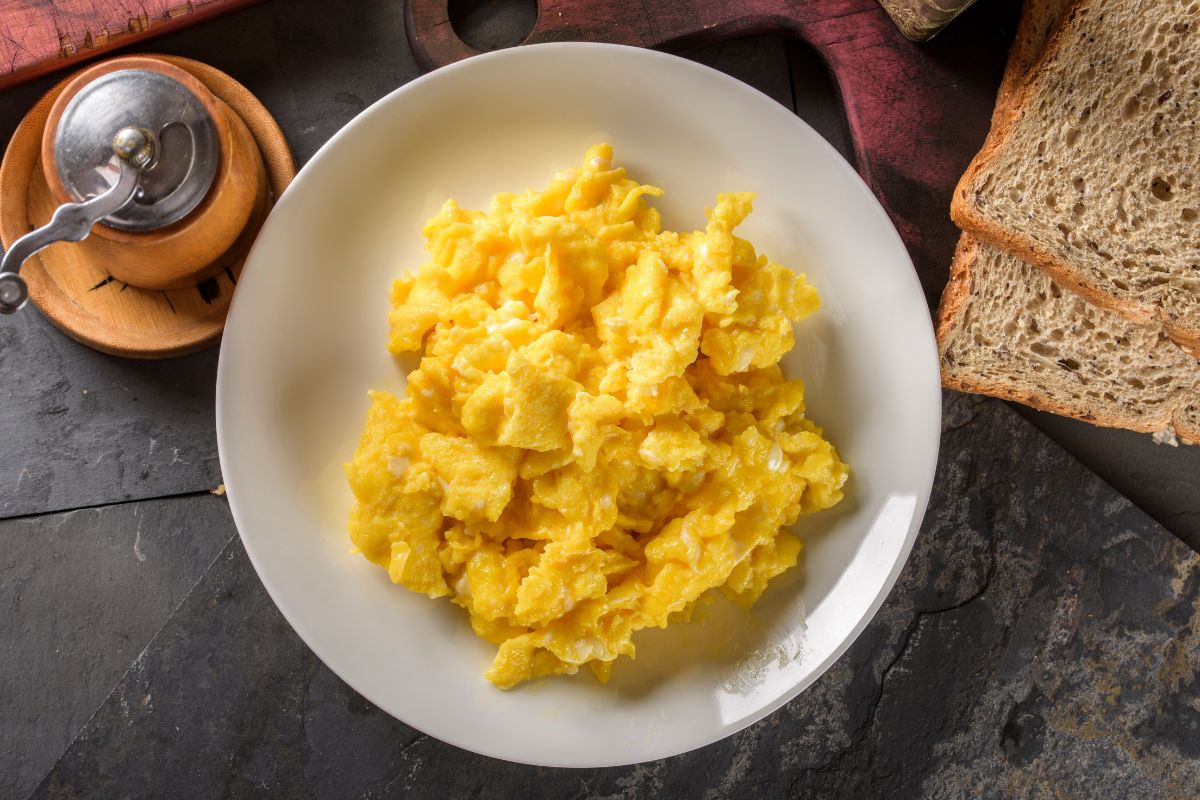 A white plate full of scrambled eggs on a black table with slices of bread.