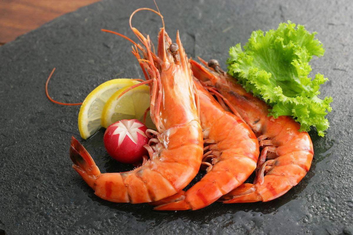 Three shrimp, sliced lemon, and vegetables on a black table.