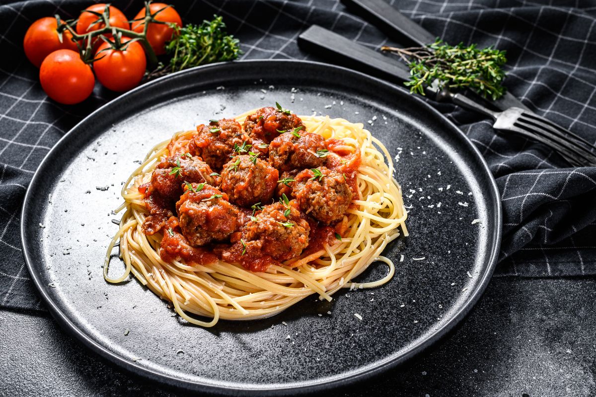 A tasty spaghetti with meatballs on a black plate with forks, herbs, and tomatoes on a table.