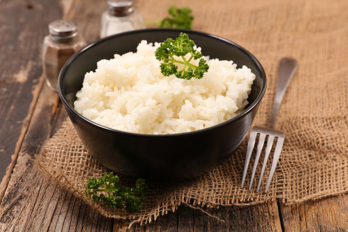 A black bowl full of cooked rice on a wooden table with a fork, salt, and pepper.