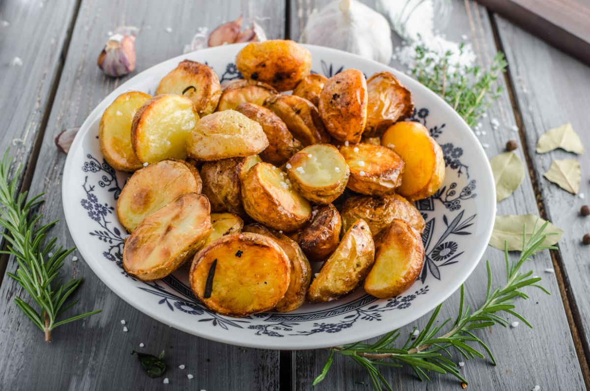 A white plate full of baked potatoes on a wooden table with garlic and herbs.