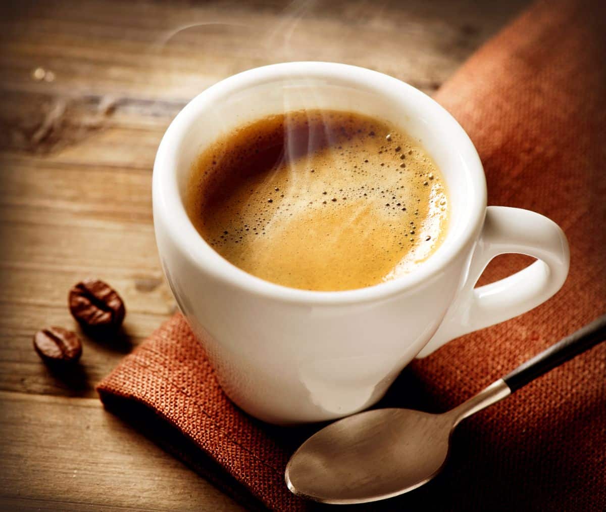 A white cup of hot coffee on a wooden table with a piece of cloth, a spoon.