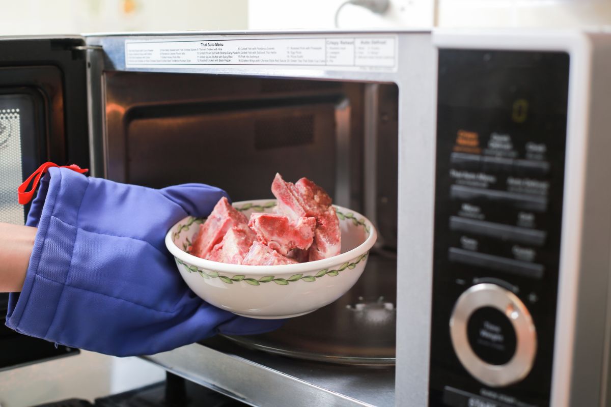 A hand with a glove putting a bowl of frozen meat in a microwave.