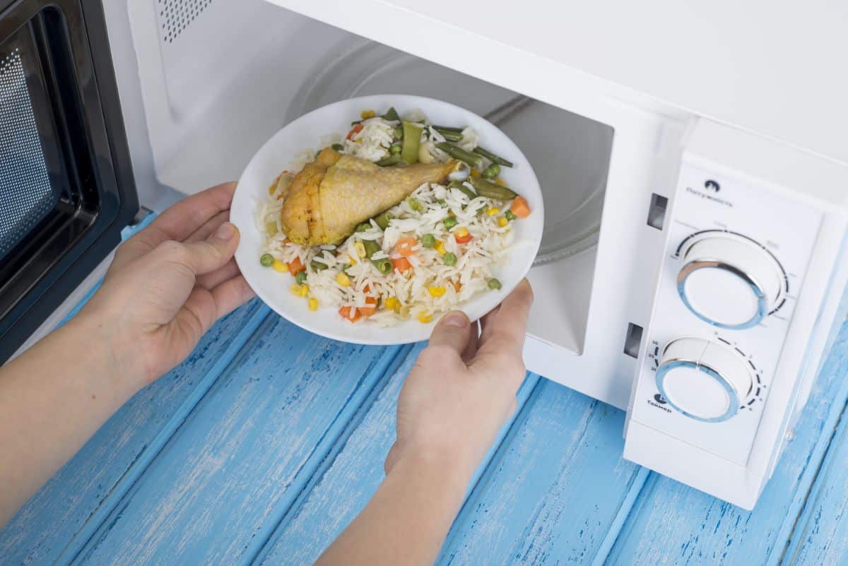 Hands putting a plate full of meal in a microwave on a blue table.