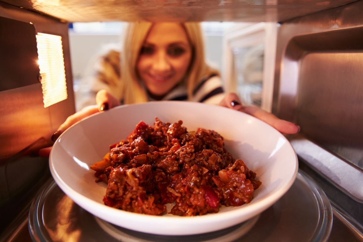 A woman putting a white plate full of leftovers in a microwave.