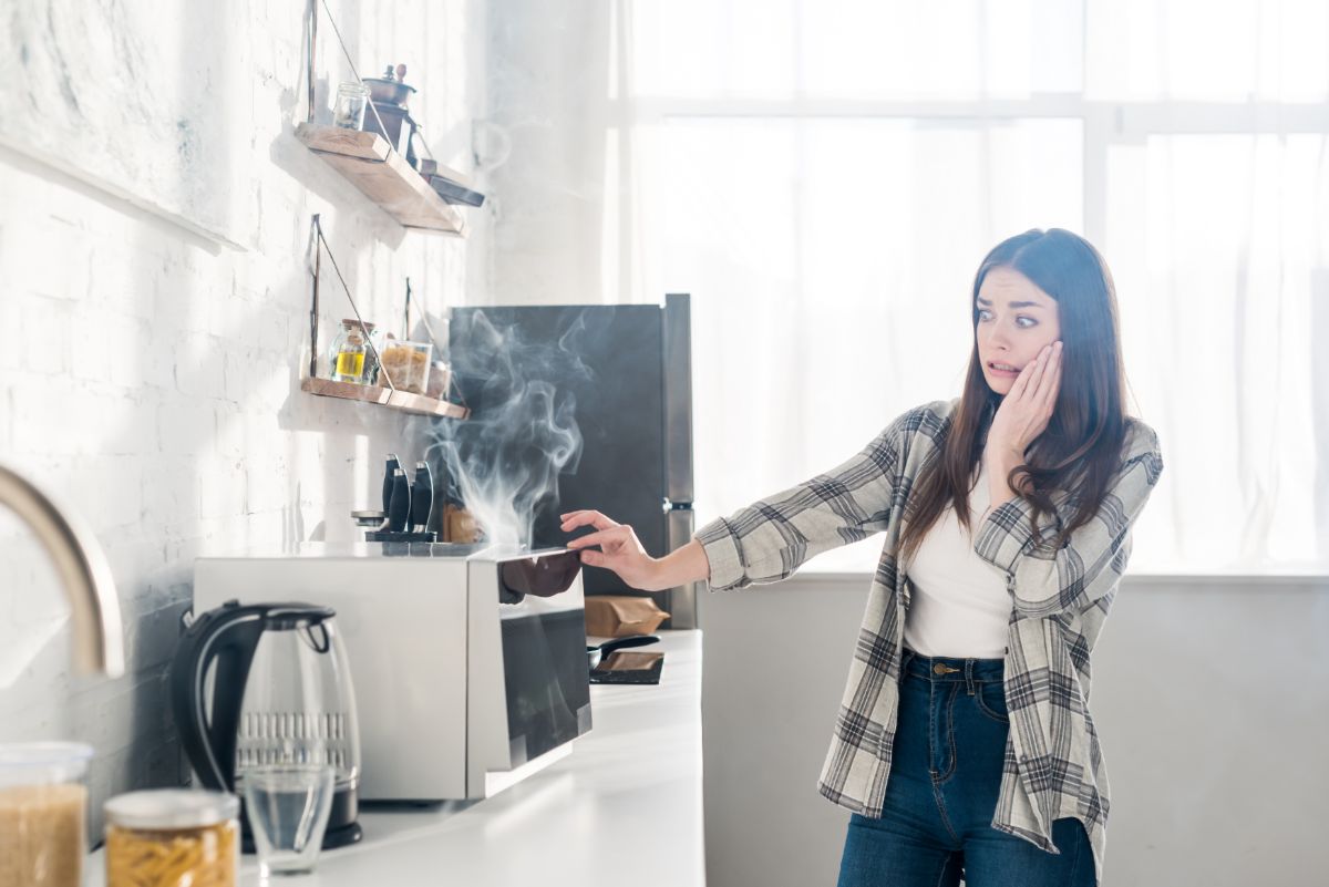 A scared young woman in a kitchen looking at a microwave.